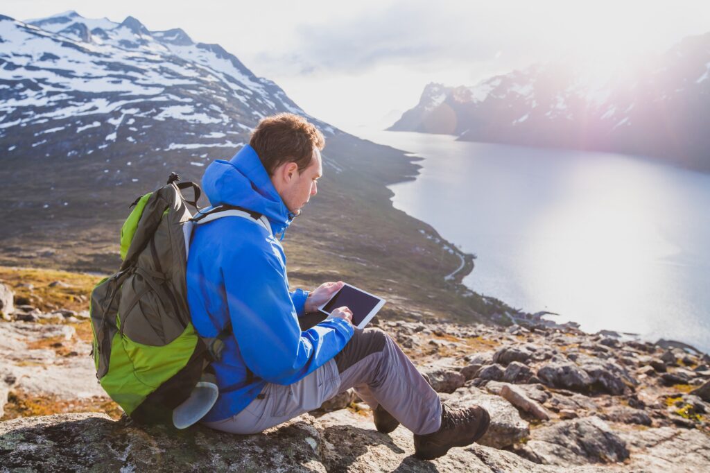 junger Tourist sitzt mit Tablet vor Bergkulisse