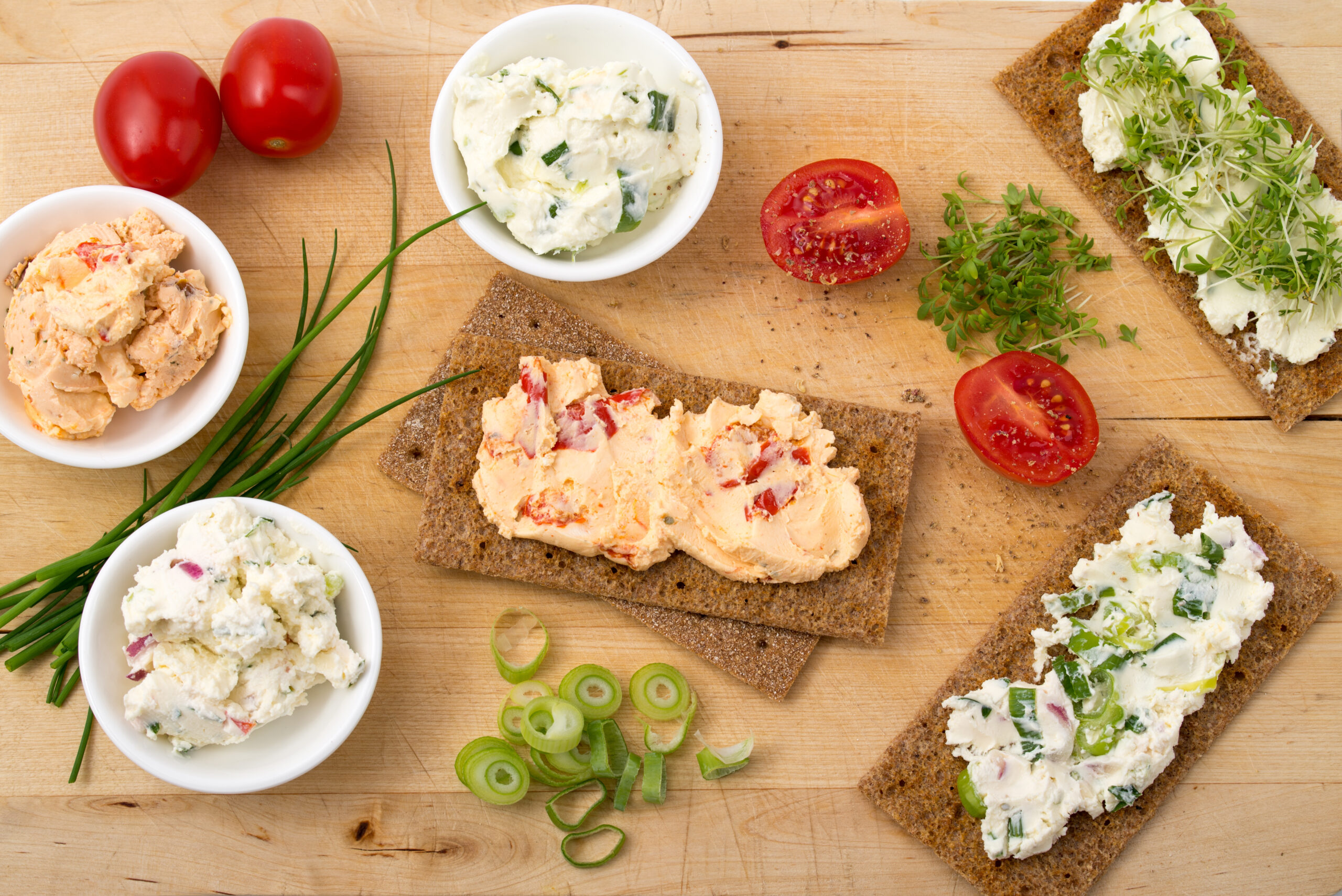 Verschiedene Kräuter- und Gewürzfrischkäse auf Broten und in Schalen, dekoriert mit Tomenten und Frühlingszwiebeln